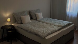 A modern bedroom with a large bed, a bedside table with a lamp, a hanging light fixture, and sheer curtains allowing natural light. - Coesfeld outskirts - Quiet apartment for 2 persons, close to town and nature (Coesfeld)