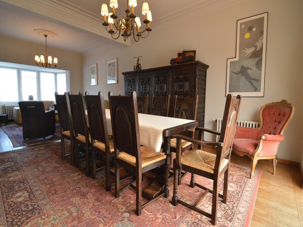 Une salle à manger avec une grande table, des chaises en bois foncé, un tapis à motifs, un lustre et un radiateur.
