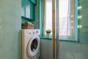 A compact laundry area with a washing machine, a wicker basket, and a potted plant by a window. - Bamboo Studio by Wonderful Italy (Turin)