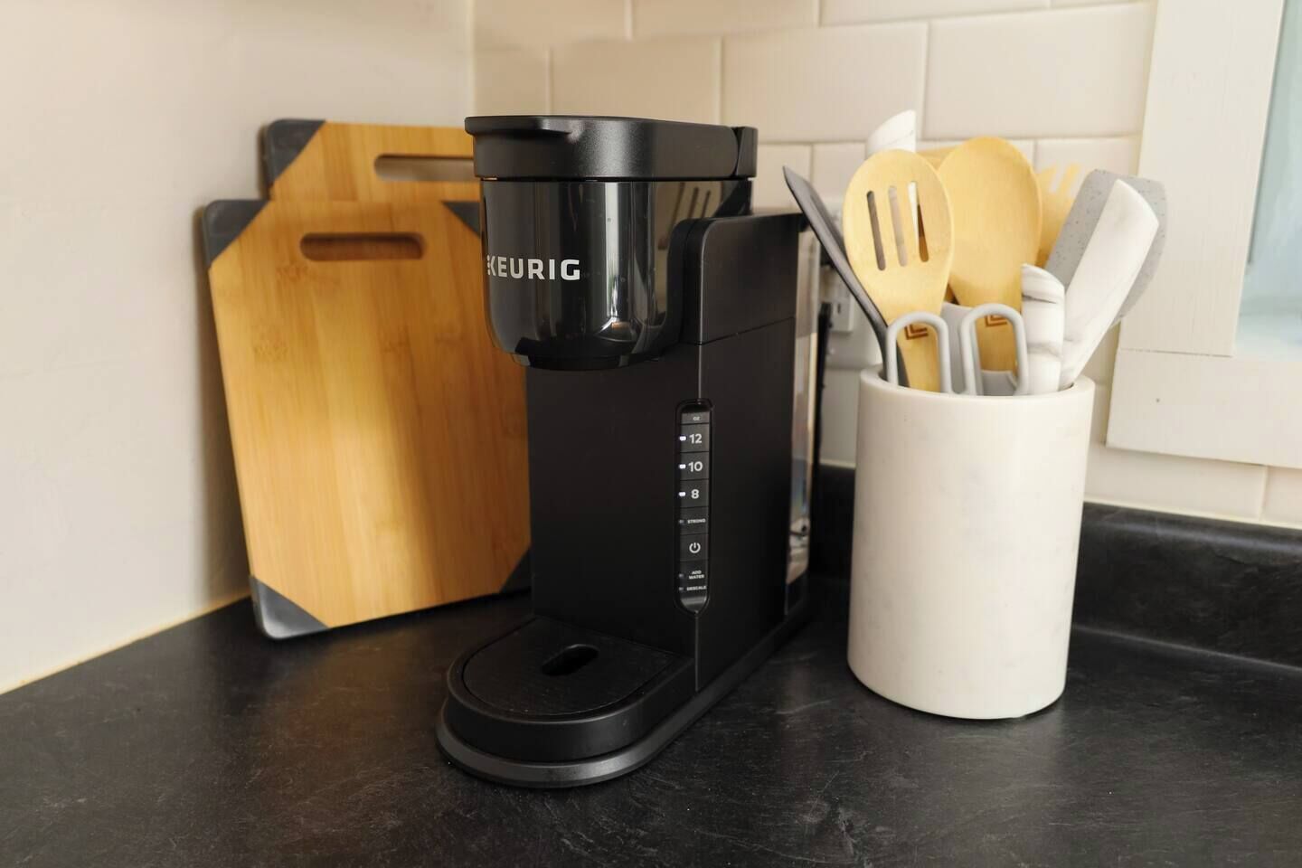 Una macchina da caffè Keurig nera su un piano di lavoro in cucina, accanto a un tagliere di legno e un portauovelli bianco con utensili di legno.