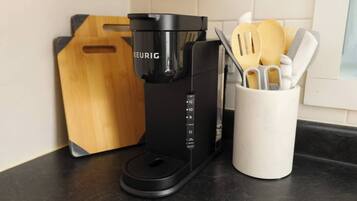Una macchina da caffè Keurig nera su un piano di lavoro in cucina, accanto a un tagliere di legno e un portauovelli bianco con utensili di legno.
