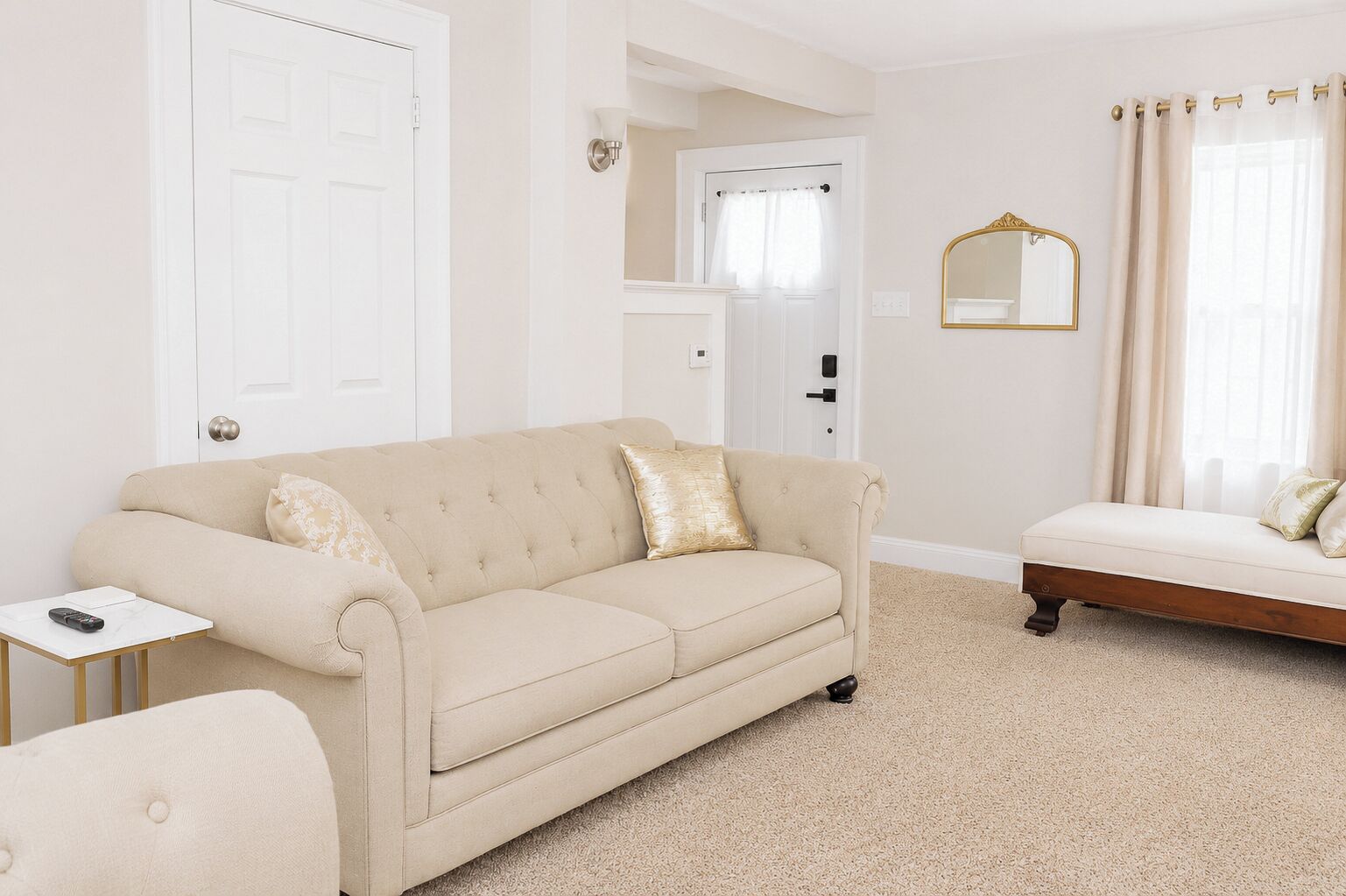 A room with a beige sofa, a small side table, a bed with a headboard, a mirror, and a door.
