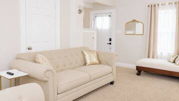 A room with a beige sofa, a small side table, a bed with a headboard, a mirror, and a door.