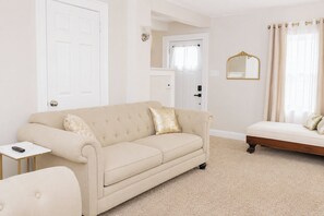 A room with a beige sofa, a small side table, a bed with a headboard, a mirror, and a door.