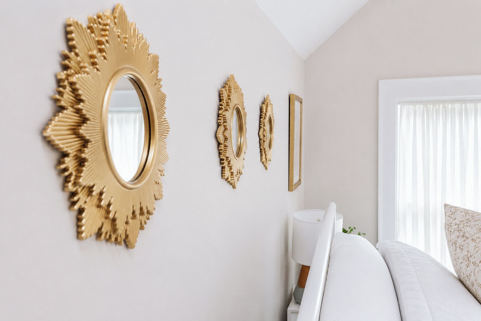 A bedroom with a large round mirror, two smaller mirrors, a bedside table, and a window with curtains.