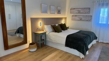 A bedroom with a bed, bedside table, mirror, ceiling fan, artwork, and a window with curtains.