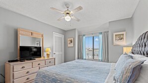 A bedroom with a bed, dresser, television, ceiling fan, and a window with curtains.