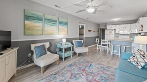 A living room with a TV, two chairs, a blue sofa, a ceiling fan, and a kitchen area with a bar.