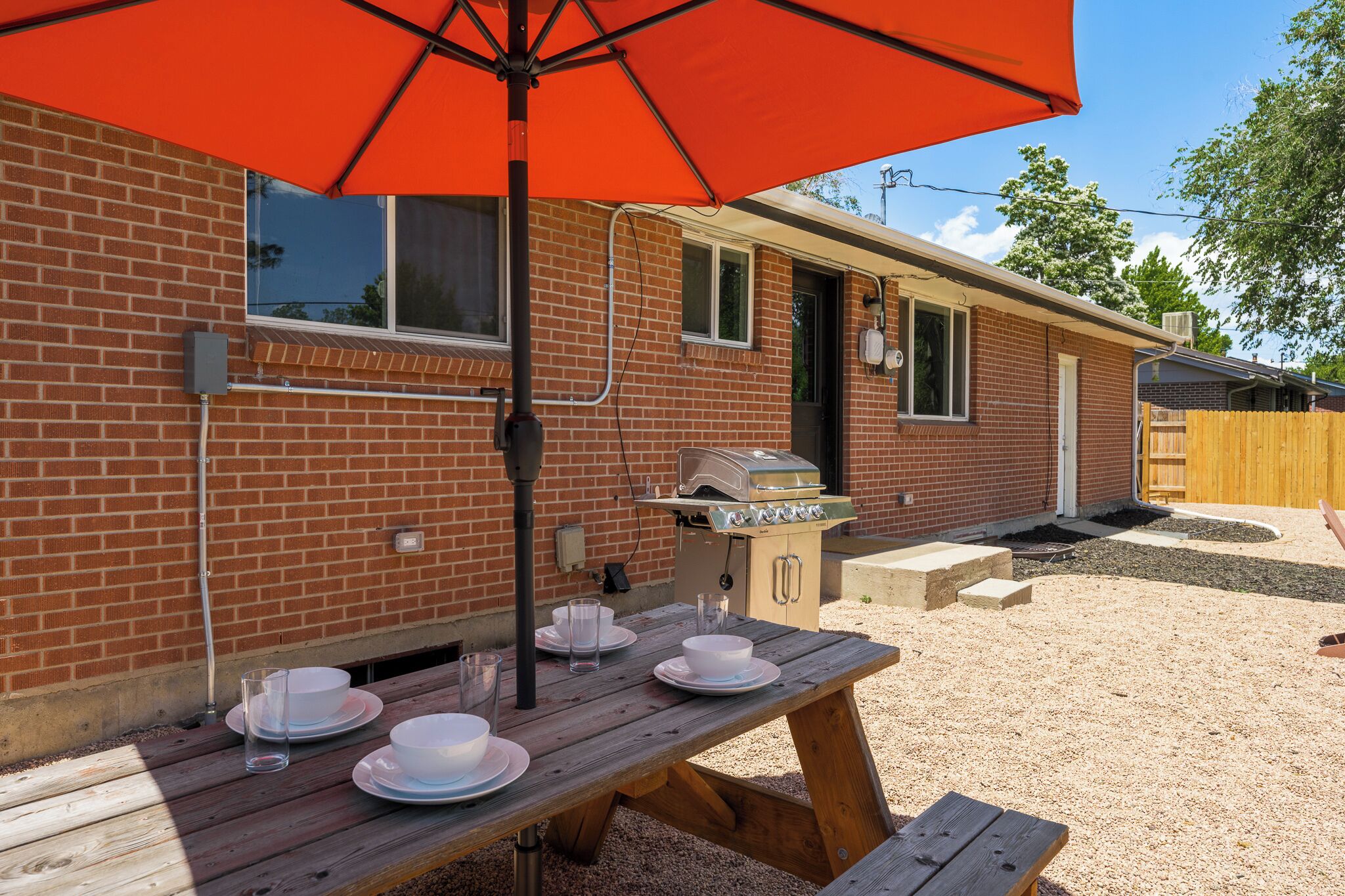 A brick house with a barbecue grill, a wooden table with plates and glasses, and a red umbrella.