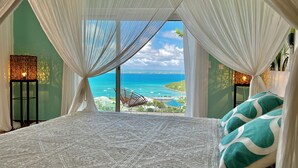 Ein Schlafzimmer mit einem großen Bett, weißen Vorhängen und Blick auf das Meer und Boote.