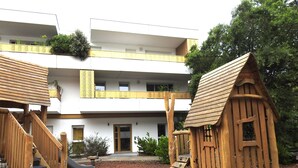 A playground with wooden structures, including a slide and climbing steps, in front of a building with a white facade and balconies.