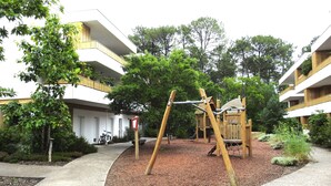 A playground with a wooden swing set and climbing structures, surrounded by trees and a building with balconies.
