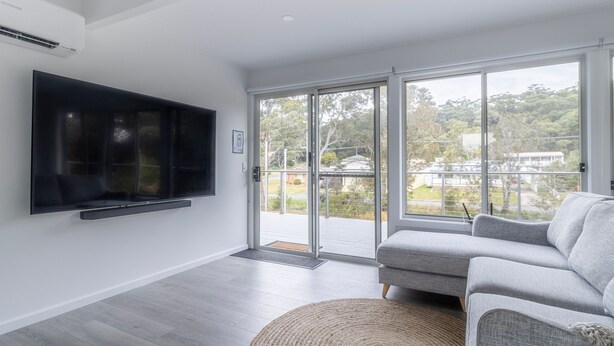 Una sala de estar moderna con un televisor de pantalla plana montado en la pared, un sofá gris y un amplio ventanal corredera que da a un balcón con vistas al vecindario.