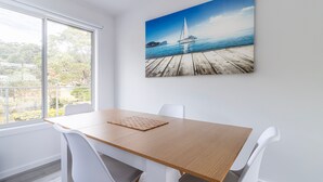 Un comedor con mesa de madera, sillas blancas y un amplio ventanal con vistas a la vegetación y un paisaje lejano.