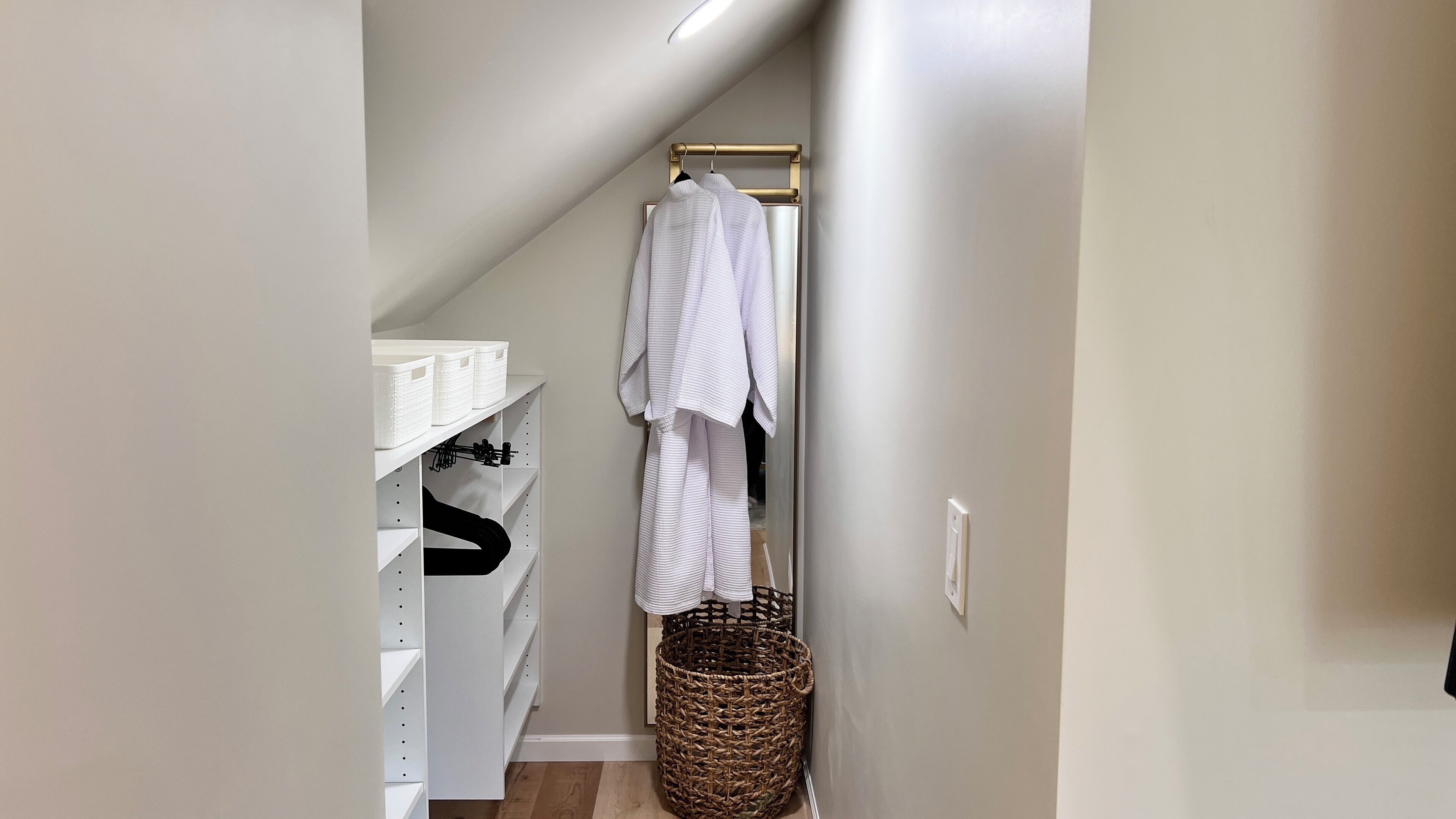 A narrow hallway with a white shelving unit, a hanging rack with clothes, and a wicker basket.