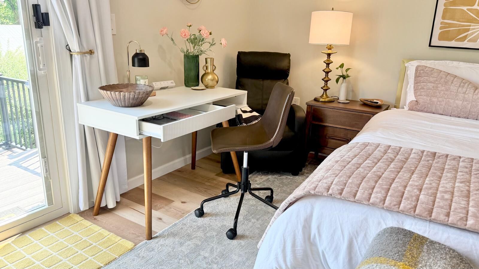 A bedroom with a bed, desk, chair, bedside table, lamp, and a window with curtains.