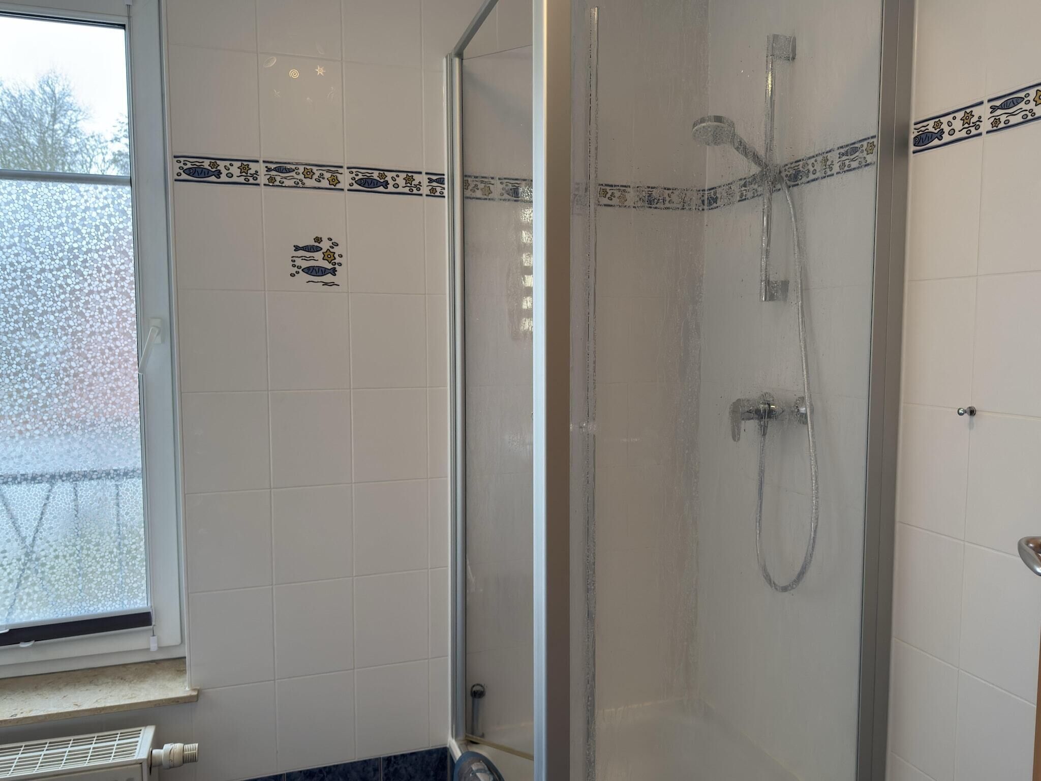 A shower with a glass door, white tiled walls, and a window with frosted glass.