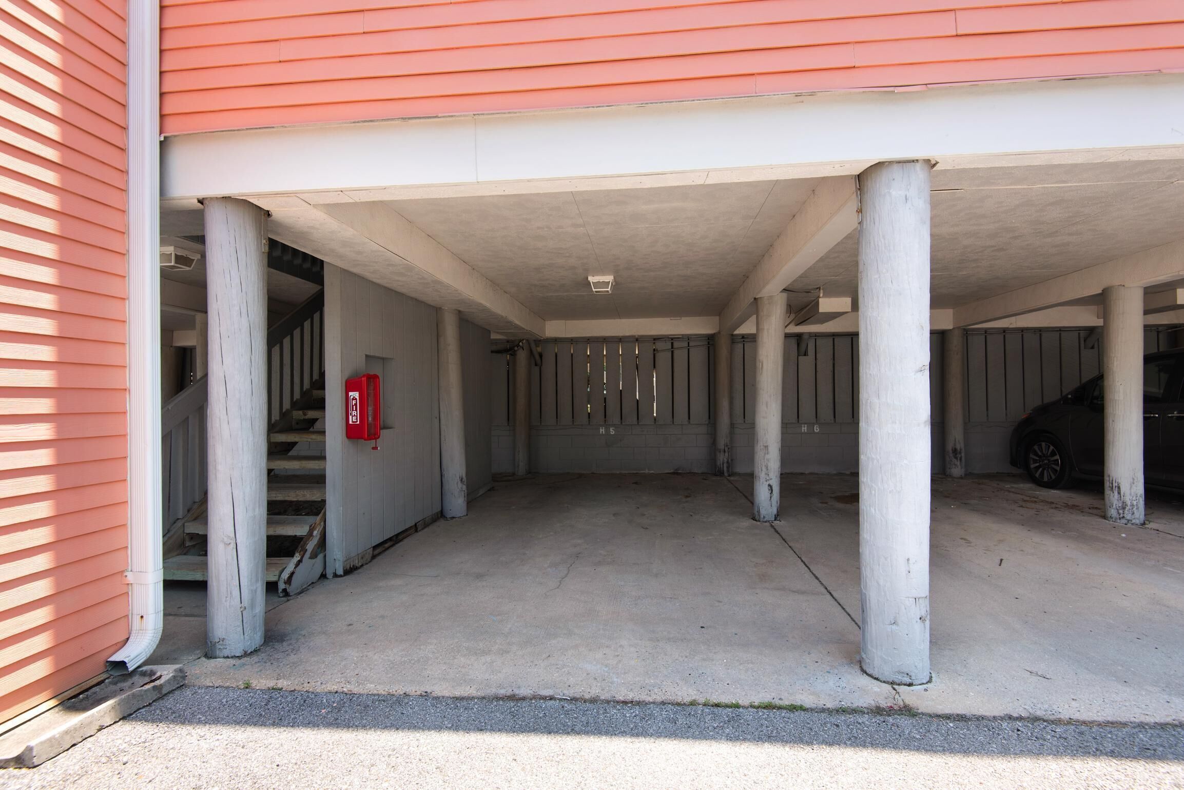A covered parking area with concrete pillars, a red fire alarm box, and a car parked to the right.