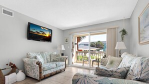 A living room with two patterned sofas, a coffee table, a TV mounted on the wall, and a sliding glass door leading to an outdoor area with a view of palm trees and a building.
