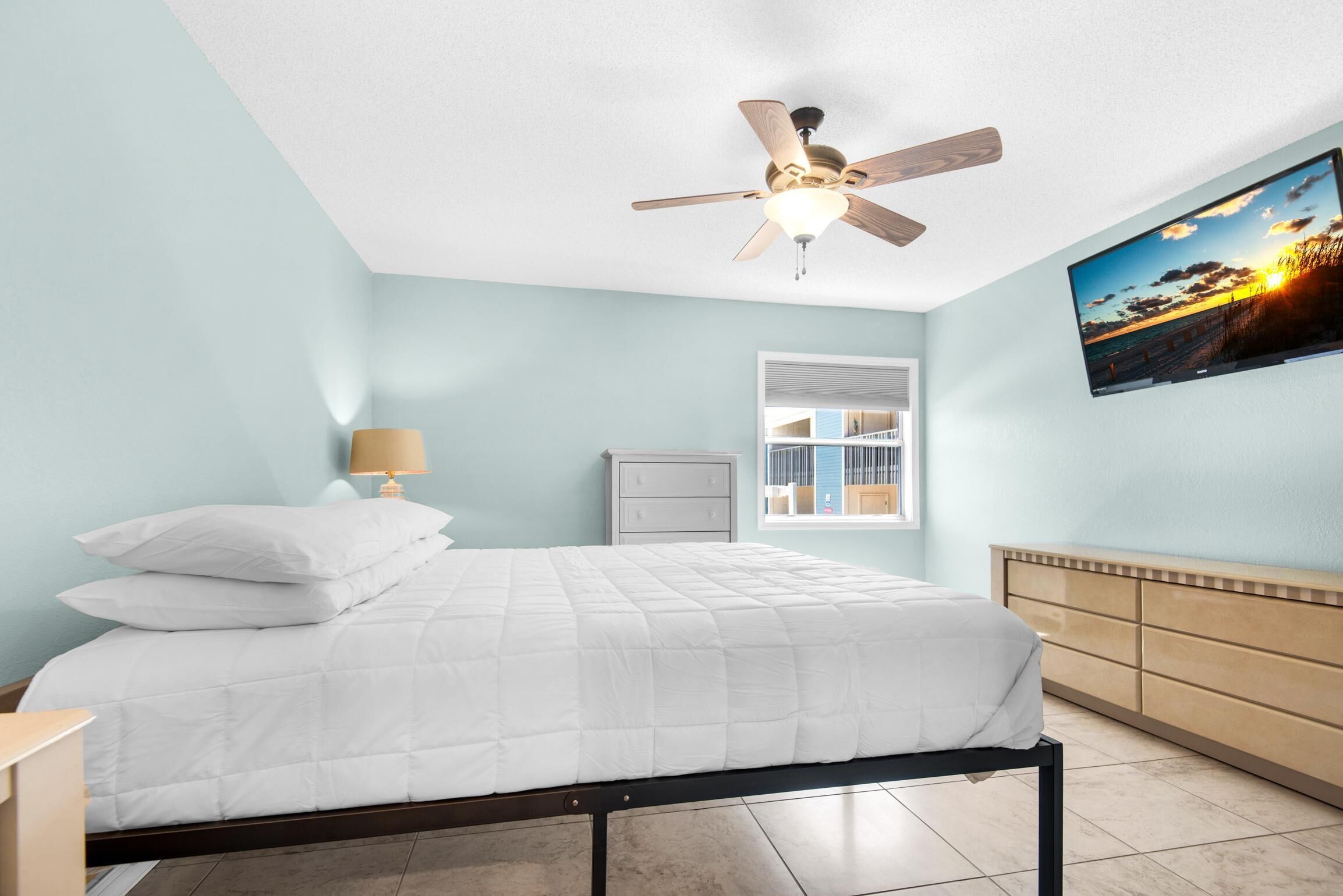 A bedroom with a bed, a ceiling fan, a TV on the wall, and a window.