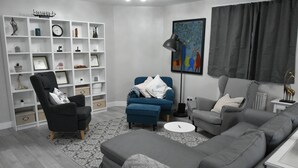 A modern living room with a grey sofa, a blue armchair, and a black armchair with a patterned throw. A white shelving unit with decorative items is against the wall. - Charming Wembley Home Near Wembley Stadium (Wembley)