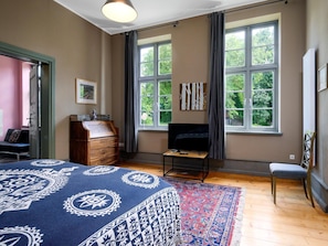 A bedroom with a bed, a TV, a desk, and a chair. There are two windows with curtains, and a door leading to another room.