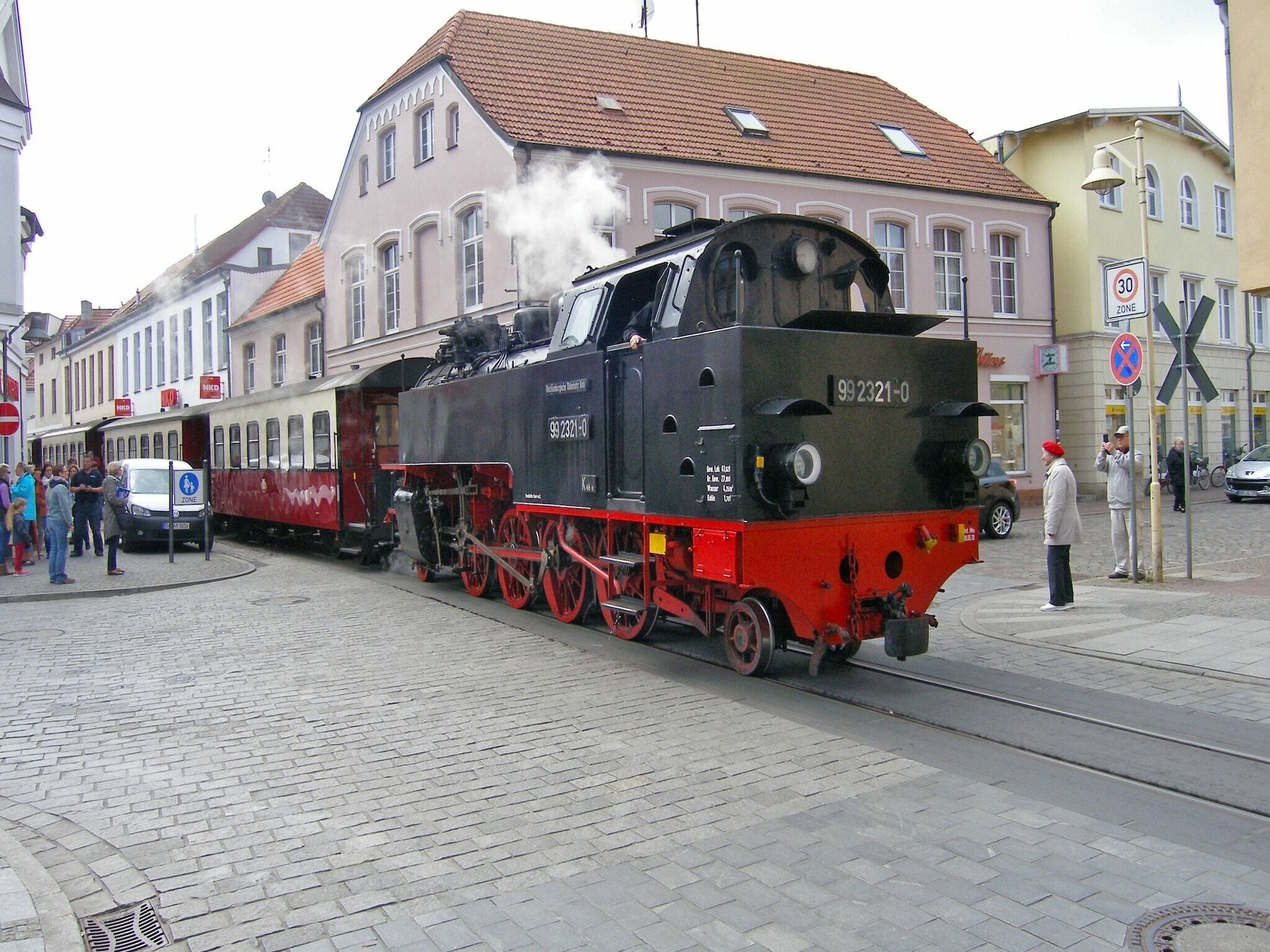 Una locomotora de vapor con el número 99 2321-0 sobre una vía, rodeada de edificios y gente.