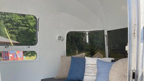 Un intérieur de camping-car compact, avec un banc rembourré de couleur grise, un tapis bleu et une fenêtre donnant sur un paysage verdoyant.