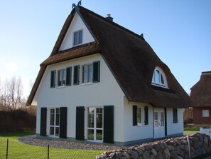 Una casa tradicional con techo de paja empinado, paredes blancas y contraventanas verdes.