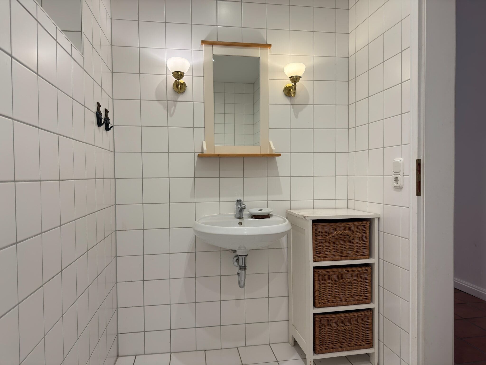 A bathroom with white tiled walls, a white sink, two wall-mounted light fixtures, a mirror, and wicker baskets on a shelf.