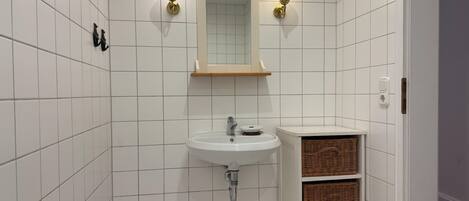 A bathroom with white tiled walls, a white sink, two wall-mounted light fixtures, a mirror, and wicker baskets on a shelf.