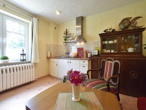 Uma cozinha com mesa de madeira, um vaso com flores, um radiador, uma janela com cortinas e um armário com portas de vidro.