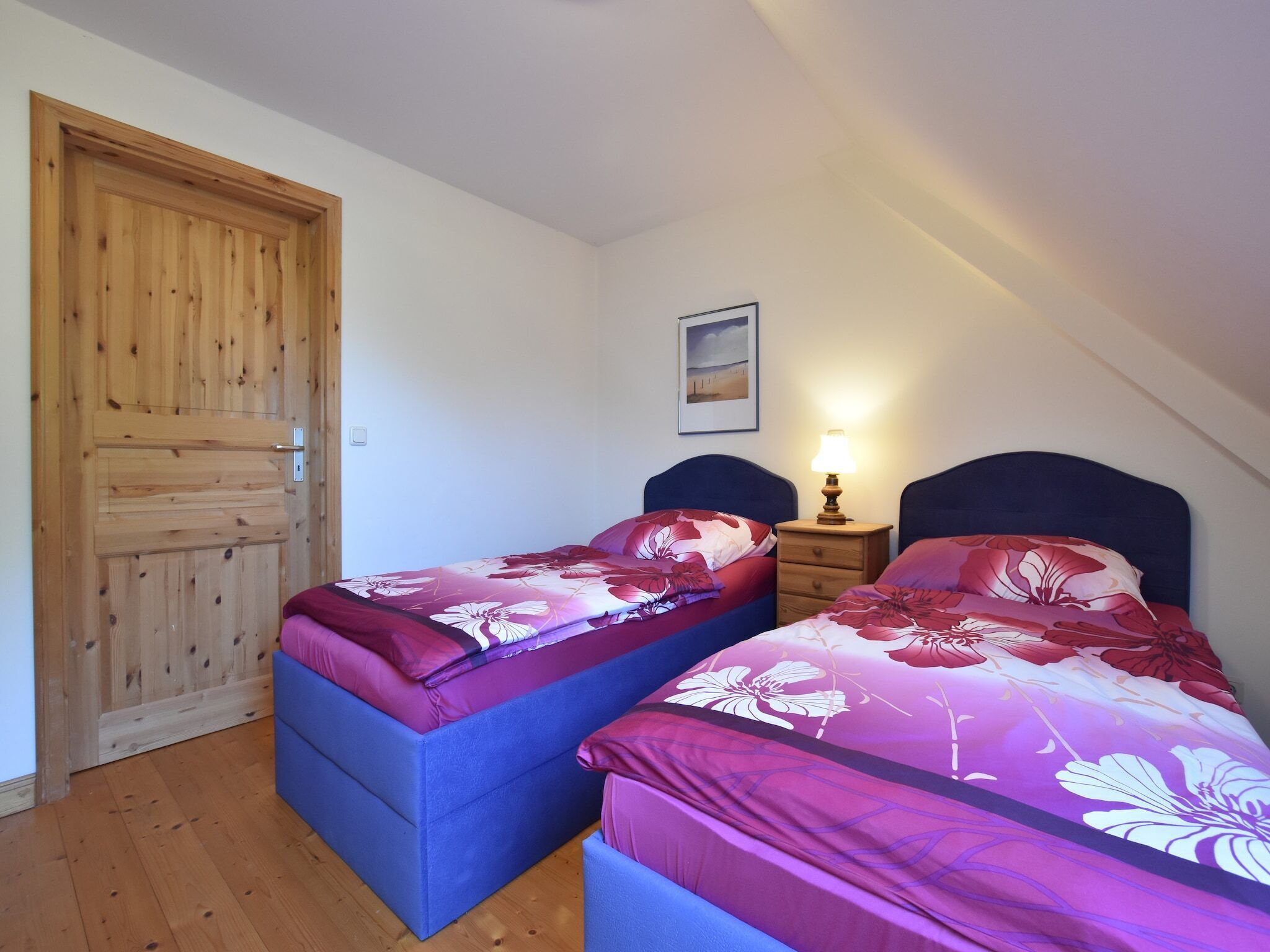 A bedroom with two beds, a wooden door, a nightstand, and a wall-mounted picture.