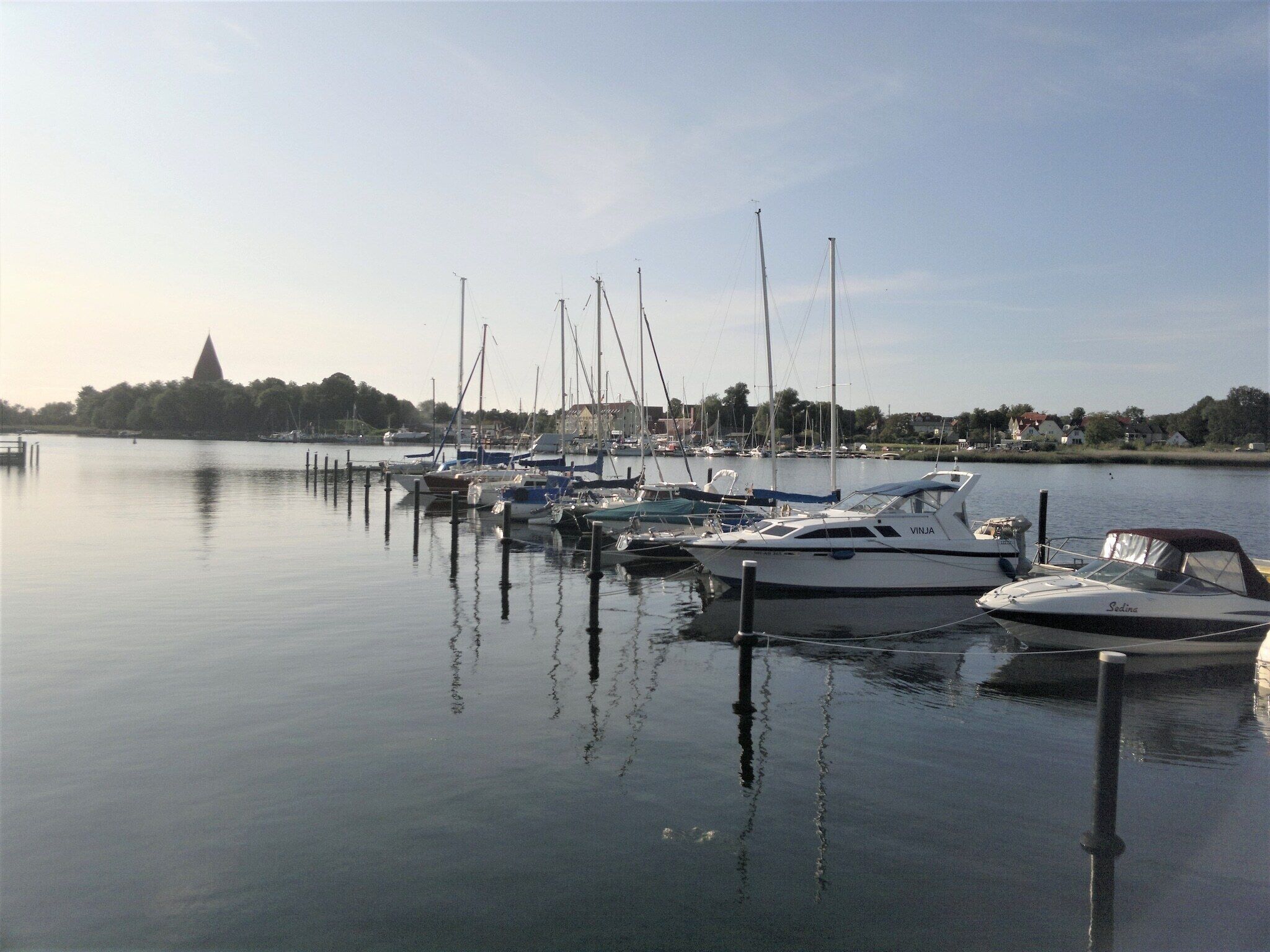Un port de plaisance où sont amarrés plusieurs bateaux, avec une eau calme et une vue lointaine sur une ville, ses maisons et son église.