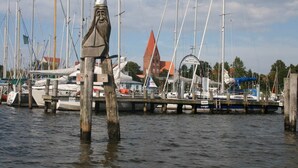 A marina with multiple boats docked, a prominent statue in the foreground, and a building with a red roof in the background. - Ferienhaus mit Kamin in Mechelsdorf (Mechelsdorf)