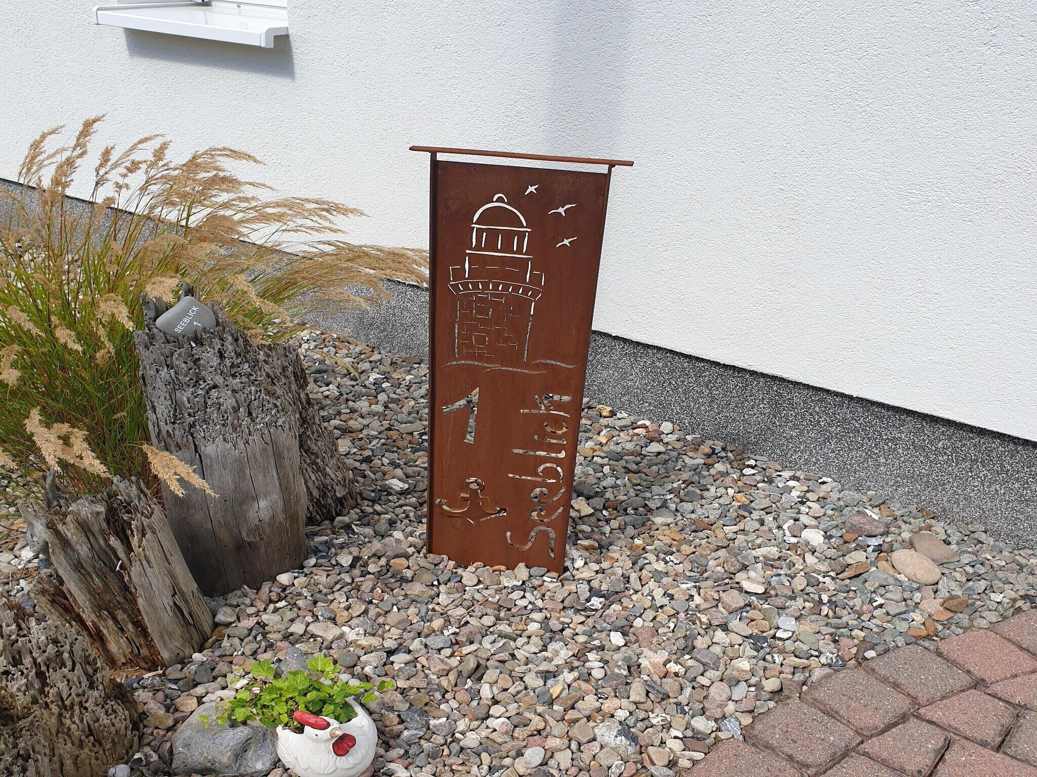 Une plaque métallique représentant un bâtiment et des oiseaux, placée sur un chemin de gravier, avec des plantes et un pot de fleurs à proximité.