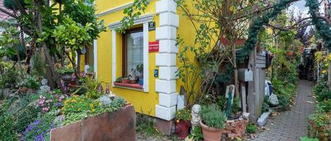 A yellow building with a small garden area in front, including a paved walkway and various potted plants.