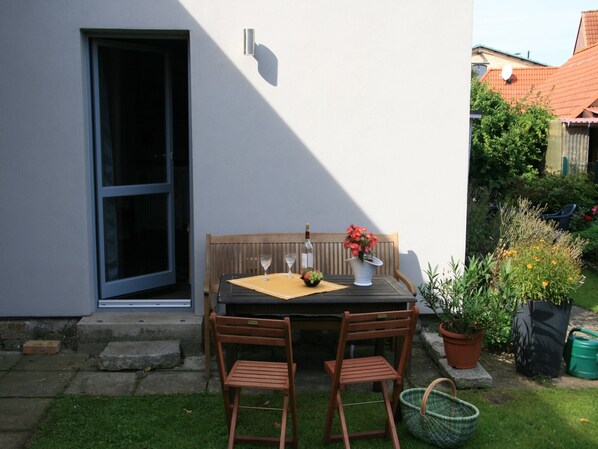 Eine Terrasse im Freien mit Tisch, Stühlen und einer Bank. Ein Blumentopf und ein Korb stehen auf dem Boden.