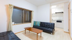 A compact living space with a sofa, a small table, a window with curtains, and a kitchenette in the background.
