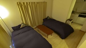 A small hotel room with two beds, a wooden bench, a small table, and a kitchenette in the background.