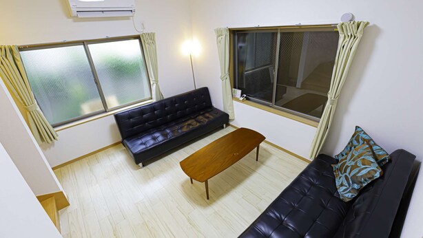 A room with a black sofa, a wooden bench, and a black leather couch. There are two windows with curtains, an air conditioning unit, and a wall-mounted speaker.