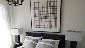 Ein ordentlich eingerichtetes Schlafzimmer mit einem Bett, zwei Nachttischen, einem modernen abstrakten Gemälde und einer Pendelleuchte.