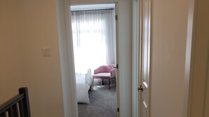 Ein Flur, der zu einem Schlafzimmer mit einem Bett, einem Sessel und einem fenster mit Vorhängen führt.