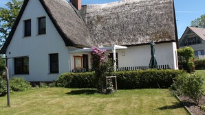 Une maison blanche avec un toit de chaume et un petit jardin agrémenté d’une plante à fleurs roses.