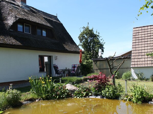 Une maison à toit de chaume, avec un jardin, un étang et une table entourée de chaises à l’extérieur.