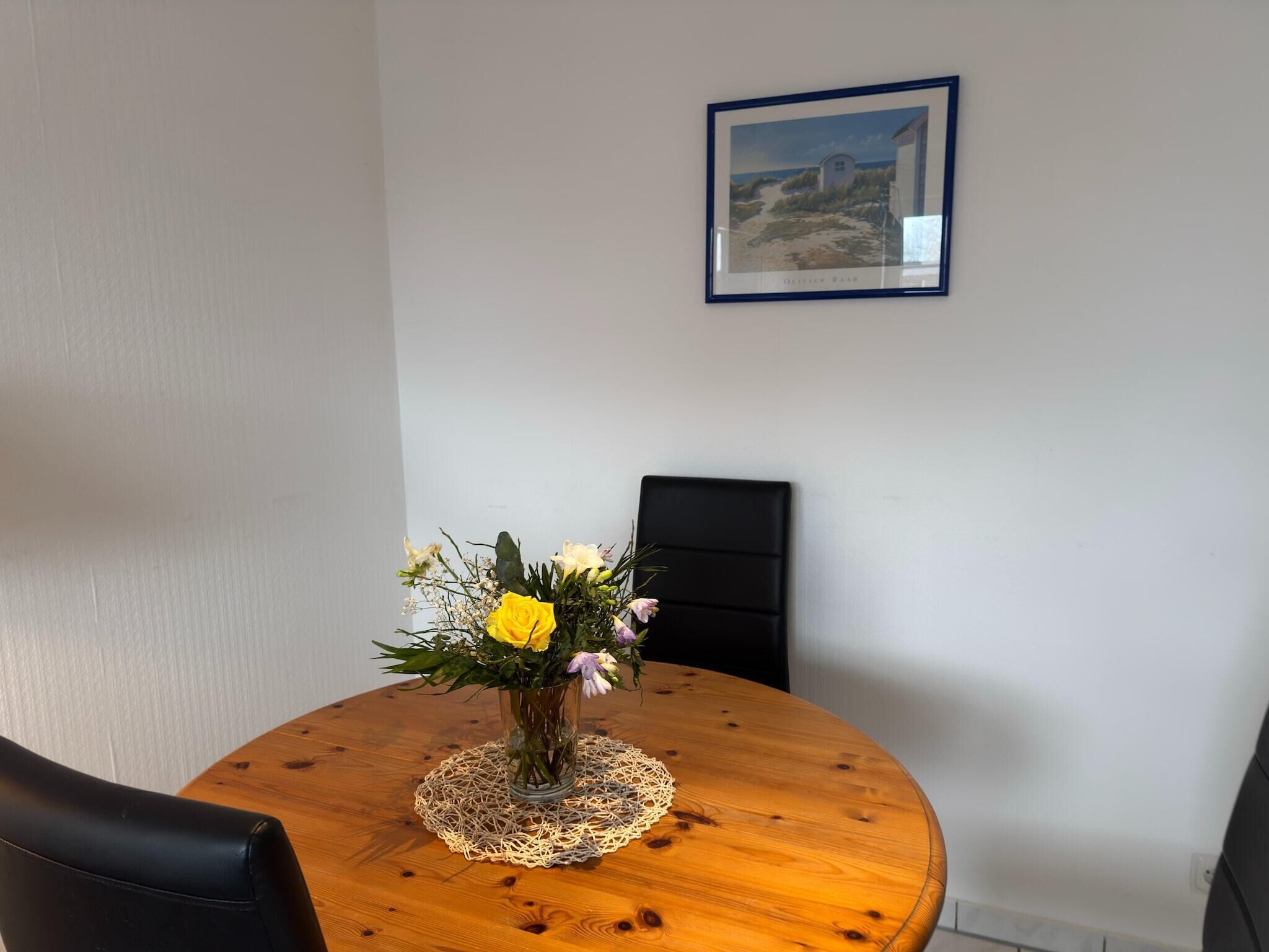 A round wooden dining table with a lace doily, a clear glass vase with flowers, and a framed picture on the wall.