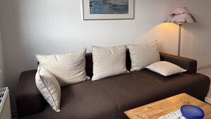 A living room with a brown sofa, three white cushions, a framed painting on the wall, a floor lamp, and a wooden coffee table with a blue bowl.
