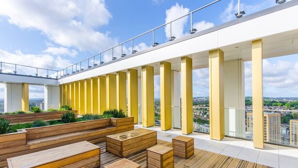 Een terras op het dak met houten zitplaatsen, gele pilaren en uitzicht over de stad.