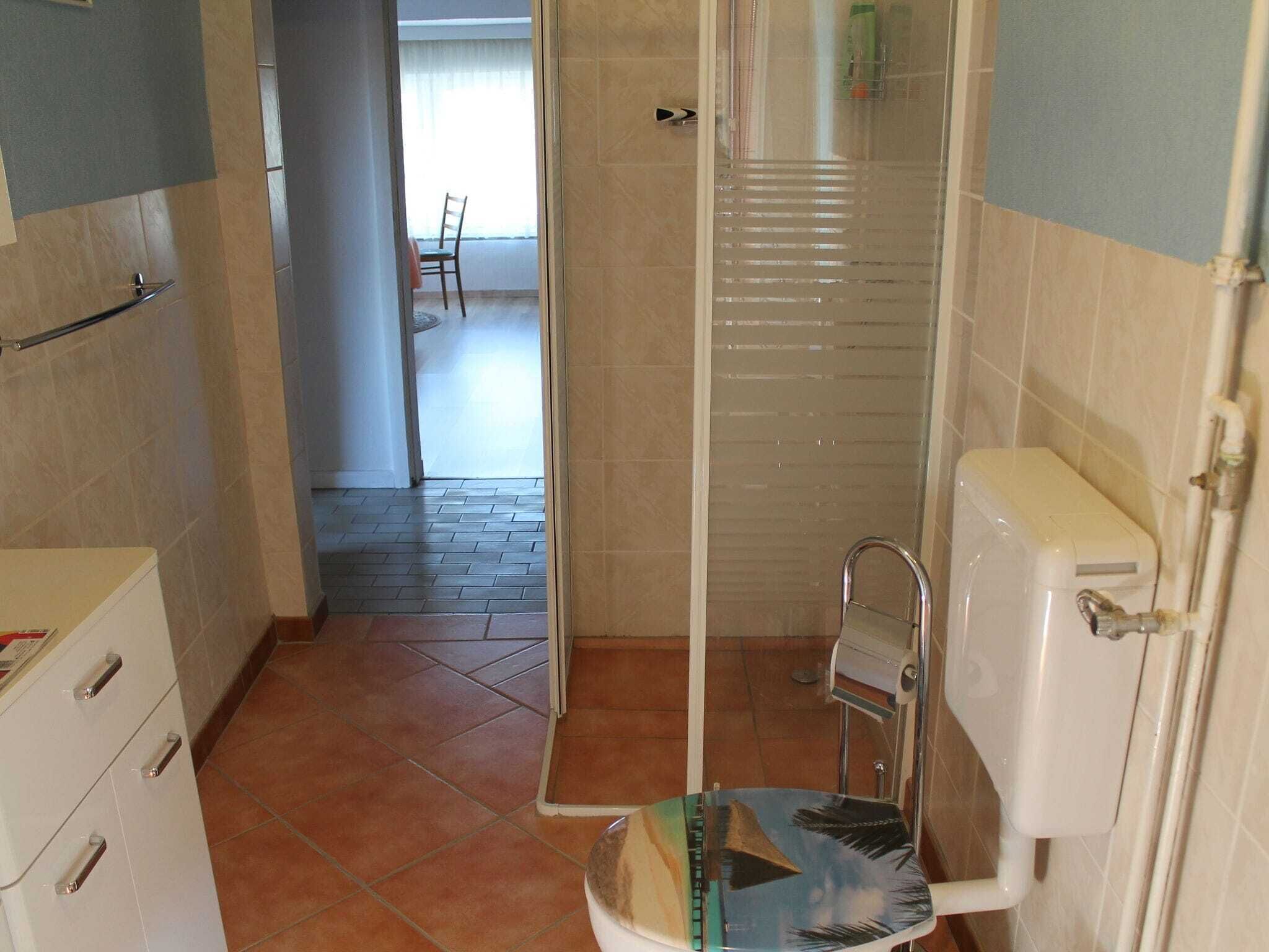 Bany amb vàter, dutxa i un petit lavabo. Hi ha una porta que dona a un passadís amb una cadira i una finestra.