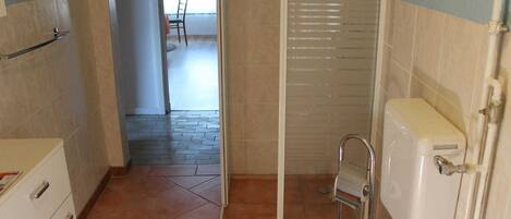 Bany amb vàter, dutxa i un petit lavabo. Hi ha una porta que dona a un passadís amb una cadira i una finestra.
